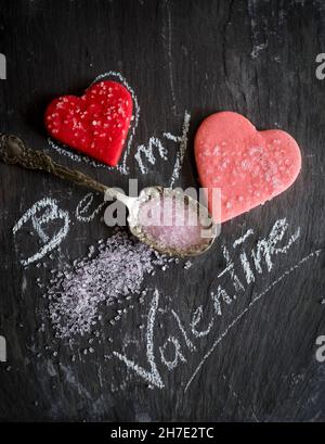 Inscription « soyez ma Saint-Valentin » à la craie avec des biscuits en forme de coeur et du sucre rose Banque D'Images