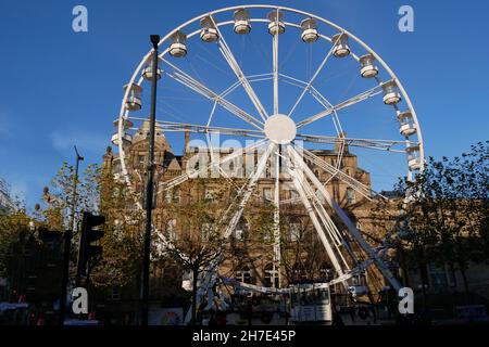 Grande roue de ferris blanche avec plusieurs gousses d'observation devant un grand bâtiment en pierre petits arbres en premier plan et ciel bleu Banque D'Images