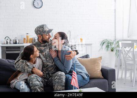 Homme en uniforme de camouflage qui embrasse la famille près du drapeau américain à la maison Banque D'Images