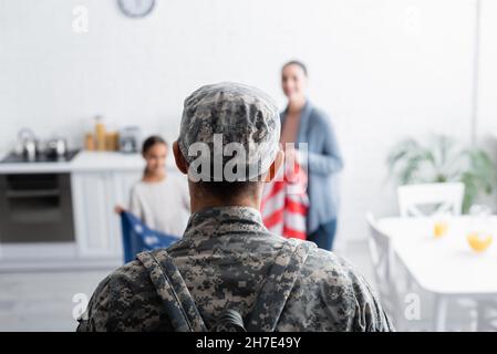 Soldat en camouflage debout près d'une famille floue avec drapeau américain à la maison Banque D'Images