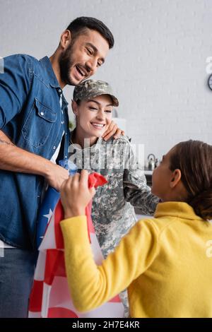 Homme souriant embrassant la femme en uniforme militaire près d'une fille avec le drapeau américain à la maison Banque D'Images