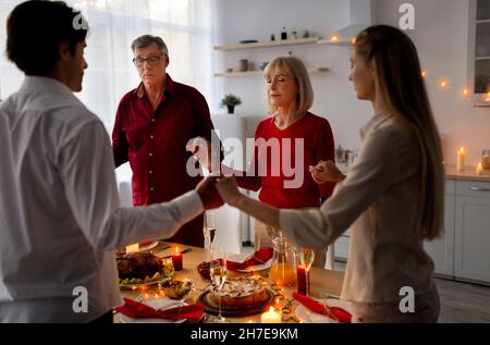 Jeune couple et parents âgés debout près de la table, tenant les mains et priant, célébrant Thanksgiving ou Noël Banque D'Images