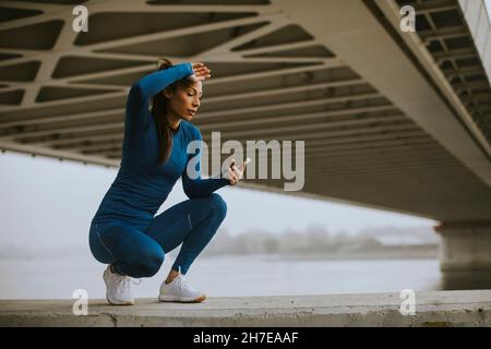 Jolie jeune femme en costume bleu de suivi de son entraînement sur l'application de fitness au bord de la rivière à l'automne matin Banque D'Images