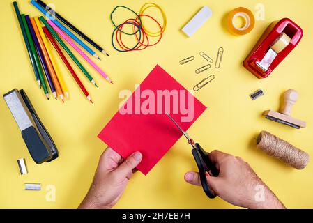 Vue de dessus des mains d'un homme coupant du papier rouge avec des ciseaux, entouré de crayons de couleur, taille-crayon, agrafeuse et divers outils d'artisanat sur un yello Banque D'Images