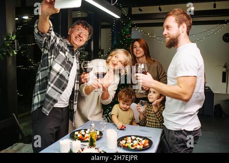 Poser pour une grande famille de selfie lors d'un rassemblement et d'une célébration de noël.Autour d'une grande table à manger, levant des verres à vin.Arbre de Noël et Banque D'Images