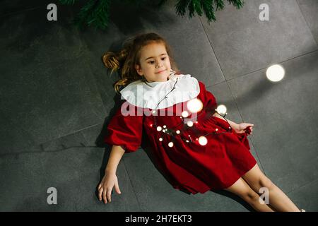 Jolie petite fille dans une robe rouge émaillée par les lumières de guirlande de noël.Recherche Banque D'Images