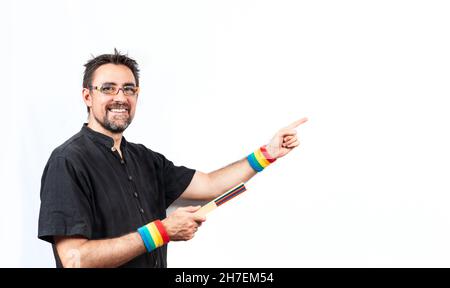 Homme en chemise noire portant des lunettes et des bracelets de couleur arc-en-ciel ; souriant à l'appareil photo et pointant vers un espace vide avec une main et un ventilateur fermé Banque D'Images