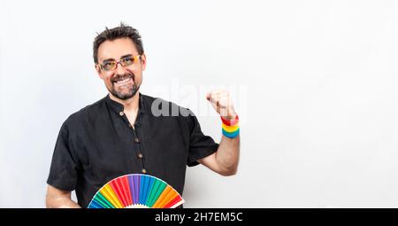 Homme aux cheveux foncés en chemise noire avec lunettes et bracelet aux couleurs de l'arc-en-ciel; avec un visage joyeux tenant un éventail arc-en-ciel ouvert et se pliant le bras serré Banque D'Images