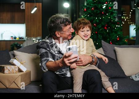 Petit-père amusant assis sur un canapé, essayant de prendre le selfie avec son petit-fils gigoleur assis sur ses genoux.Arbre de Noël en arrière-plan. Banque D'Images