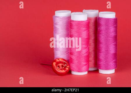 Quatre bobines de fil à coudre de différentes nuances de rose avec un bouton rouge et une aiguille isolée sur fond rouge Banque D'Images