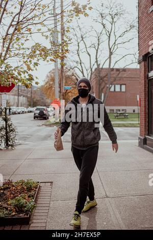 Un jeune homme de course mixte commande au comptoir du café de quartier portant un masque noir et un bonnet.Dans le café portant un masque et de social distancer pendant COVID Banque D'Images