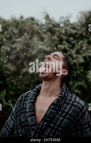 Un jeune homme de course mixte mange des flocons de neige tout en dansant dans la première neige de la saison pendant qu'il est dans une chaise longue intérieure, robe en flanelle. Banque D'Images