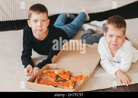 deux garçons se trouvent sur le sol en mangeant de la pizza pepperoni dans une boîte. cadre vertical. deux garçons se trouvent sur le sol en mangeant de la pizza pepperoni dans une boîte. Banque D'Images