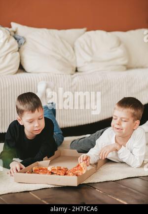 deux garçons se trouvent sur le sol en mangeant de la pizza pepperoni dans une boîte. cadre vertical. deux garçons se trouvent sur le sol en mangeant de la pizza pepperoni dans une boîte. Banque D'Images