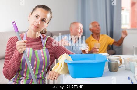 Femme fatiguée pendant les travaux ménagers, homme âgé avec homme regardant la télévision Banque D'Images