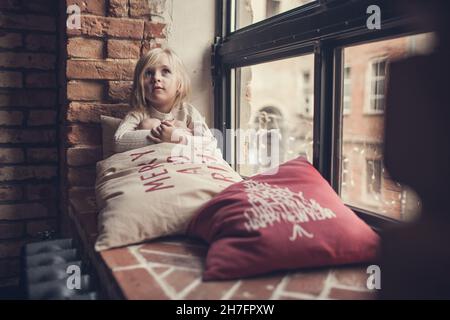 Une petite fille est assise sur un rebord de fenêtre en brique et soutient sa tête avec sa main entourée d'oreillers avec des inscriptions de Noël et fait des souhaits à San Banque D'Images