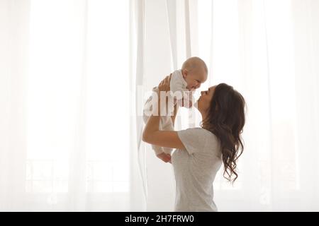 Bonne mère souriante avec bébé s'amusant ensemble sur blanc des rideaux de l'arrière-plan de la fenêtre Banque D'Images