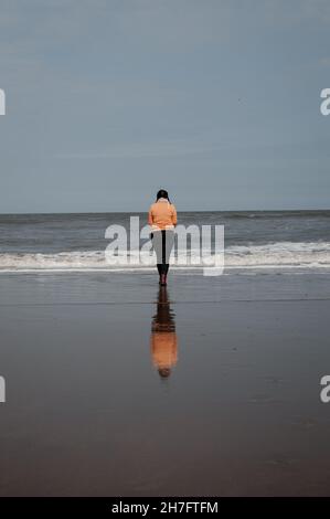 Une figure solitaire se tient au bord de l'océan, sa réflexion se jette sur le sable Banque D'Images