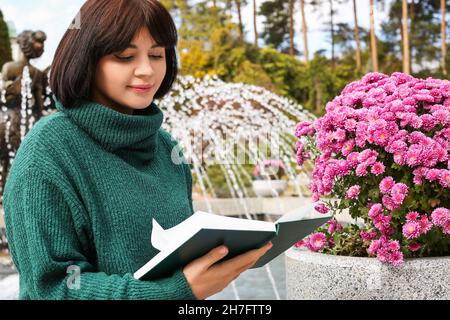 Jeune femme lisant le livre près du parterre de fleurs avec de belles fleurs dans le parc d'automne Banque D'Images