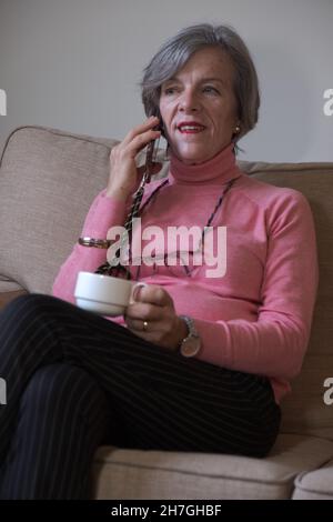 Bonne femme mûre parlant téléphone cellulaire tout en étant assise sur le canapé avoir le café Banque D'Images