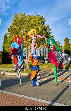 Ensemble complet de robes à tête courbettes avec visages peints en costumes colorés tenant les mains et s'amusant en se tenant debout sur des pilotis dans le parc, le jour ensoleillé Banque D'Images