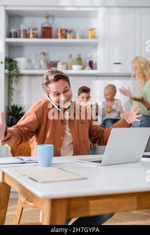 homme souriant qui geste tout en parlant sur un smartphone près d'un ordinateur portable et de la famille brouillée boire du jus d'orange dans la cuisine Banque D'Images
