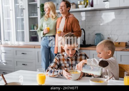 garçon versant du lait dans des flocons de maïs tout en breakfasting avec frère près de la famille floue Banque D'Images