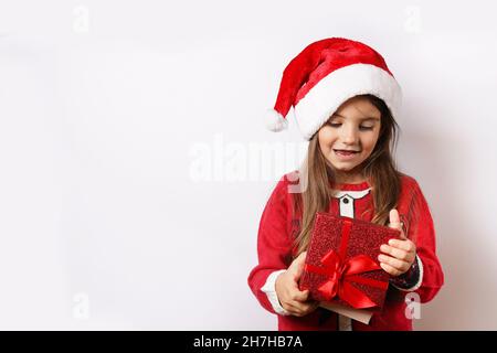 Joyeux enfant fille dans un chapeau de Noël tenant une boîte cadeau rouge isolée sur fond blanc Banque D'Images