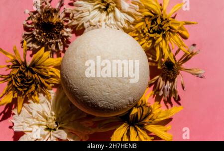 Bombe de bain blanche parmi les bourgeons de fleurs séchées sur fond rose. Banque D'Images