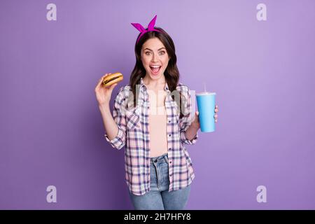 Portrait d'une fille gaie attrayante buvant du soda mangeant savoureux hamburger de bœuf délicieux isolé sur fond violet vif de couleur violet Banque D'Images