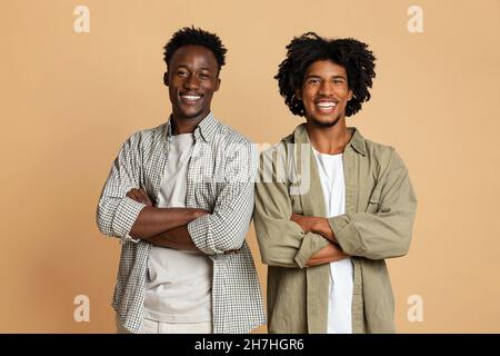 Deux amis joyeux de sexe masculin noir posant avec des bras pliés sur un arrière-plan beige Banque D'Images