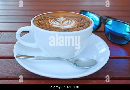 Gros plan sur une tasse de café Cappuccino chaud avec des lunettes de soleil floues en arrière-plan Banque D'Images