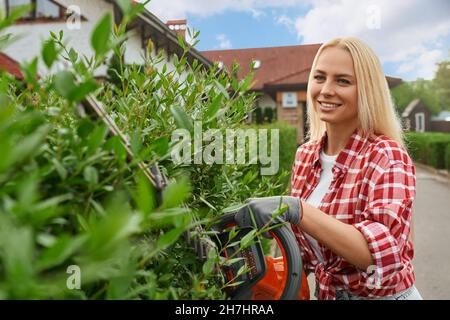 Belle jeune femme avec cheveux blonds façonnant des buissons avec machine à couper l'essence.Jardinier professionnel féminin prenant soin des plantes vertes sur le jardin arrière. Banque D'Images