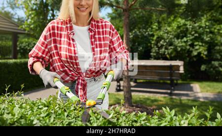 Gros plan de la femme caucasienne avec des cheveux blonds en utilisant des ciseaux de coupe tout en travaillant avec des plantes sur la cour.Jardinière féminine façonnant des buissons trop grands à l'extérieur. Banque D'Images