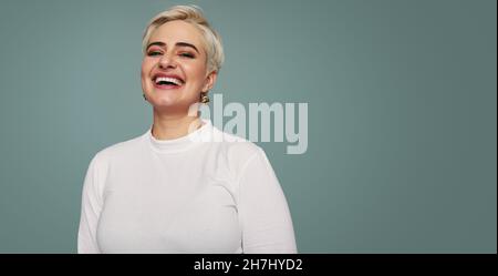 Femme blonde souriante debout seule dans un studio.Bonne femme mi-adulte regardant l'appareil photo joyeusement en se tenant debout contre un arrière-plan de studio. Banque D'Images