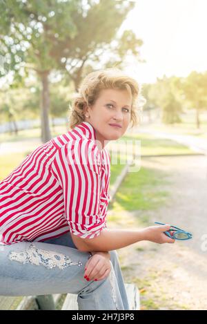 Femme blonde d'âge moyen assise sur un banc de parc tenant des lunettes de soleil et regardant la caméra Banque D'Images