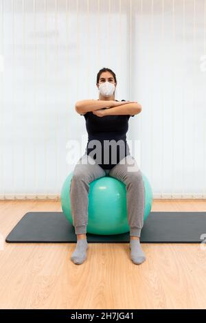 femme enceinte portant un masque facial faisant des exercices pilates assis sur une balle de fitness avec ses bras devant Banque D'Images