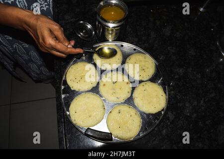 Femme cuisine à la main faisant populaire sud indien plat à la vapeur rava idli ou semolina idli. Verser le ghee ou le beurre clariné sur la préparation de la cuisine de l'idlis Banque D'Images