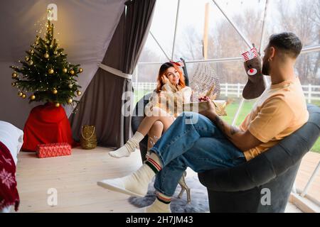 Un jeune couple est assis dans un confortable glamping.L'homme donne un cadeau à la femme pour Noël. Banque D'Images