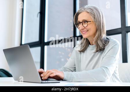 Femme d'âge moyen assise sur le bureau et bavardant en ligne sur l'ordinateur portable.Joyeuse femme d'affaires portant des lunettes et décontracté de taper sur le clavier, regarde l'écran satisfait Banque D'Images