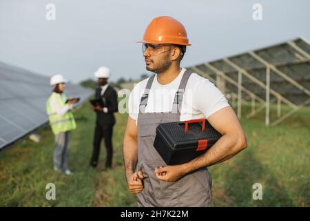 Homme indien dans une combinaison, des lunettes et un casque orange tenant des instruments et regardant de côté en se tenant au milieu de la station solaire.Groupe d'ingénieurs ayant discuté sur les antécédents. Banque D'Images