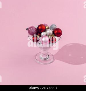 Un verre de martini rempli de boules de Noël rouge brillant, rose, blanc et gris sur fond rose.Concept tendance minimaliste pour les cartes de fête ou les bann Banque D'Images