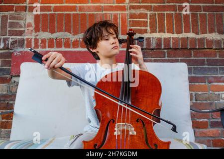 Un garçon sérieux joue du violoncelle à l'extérieur contre un mur de briques Banque D'Images