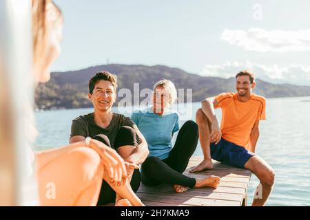 Des amis masculins et féminins parlent les uns aux autres tout en étant assis sur la jetée au bord du lac Banque D'Images
