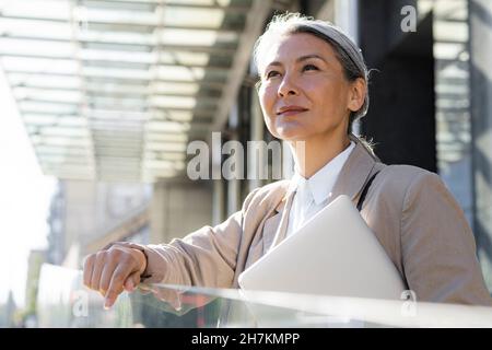 Femme d'affaires attentionnée avec ordinateur portable debout près d'une rambarde en verre Banque D'Images