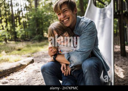 Père embrassant son fils assis sur un toboggan dans la forêt Banque D'Images