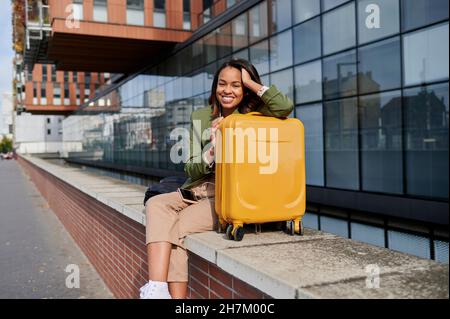 Jeune femme souriante avec la main dans les cheveux penchée sur les bagages Banque D'Images