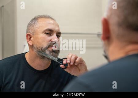 Portrait d'un beau homme mature qui tond sa barbe debout dans la salle de bains Banque D'Images