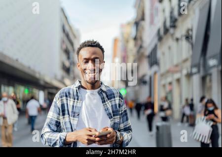 Homme souriant portant une chemise à carreaux tenant un smartphone en ville Banque D'Images