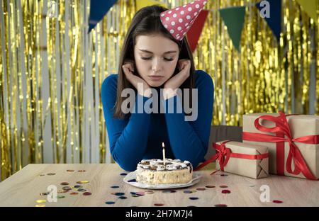 triste fille célèbre les vacances, elle est seule à la table avec un gâteau et des bougies.Casquette de fête sur la tête.Concept d'anniversaire ennuyeux. Banque D'Images